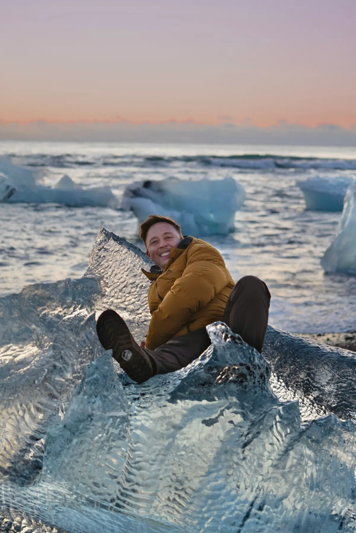 Justin grinning in a mustard puffer jacket, sitting on a block of glacier ice by the sea at sunset